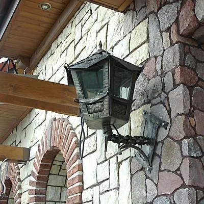 Kuta latarnia elewacyjna wykonana technikami metaloplastycznymi z ozdobną plecionką, rozetą oraz fakturowanymi detalami, przeznaczona do kamiennych rezydencji w stylu rustykalnym.