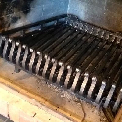 Kuty ruszt kominkowy 20 prętów 16 × 16 mm do trapezowego paleniska, ze wzmocnieniami od spodu, kutymi nóżkami i profilowanymi końcówkami.