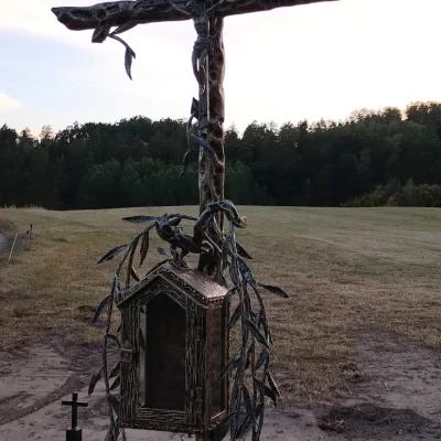 Artystyczny krzyż przydrożny w formie drzewa z wotywną kapliczką i gałązkami wierzby osadzony na kamieniu, wykonany na zamówienie Urzędu Miasta i Gminy w Mrągowie.